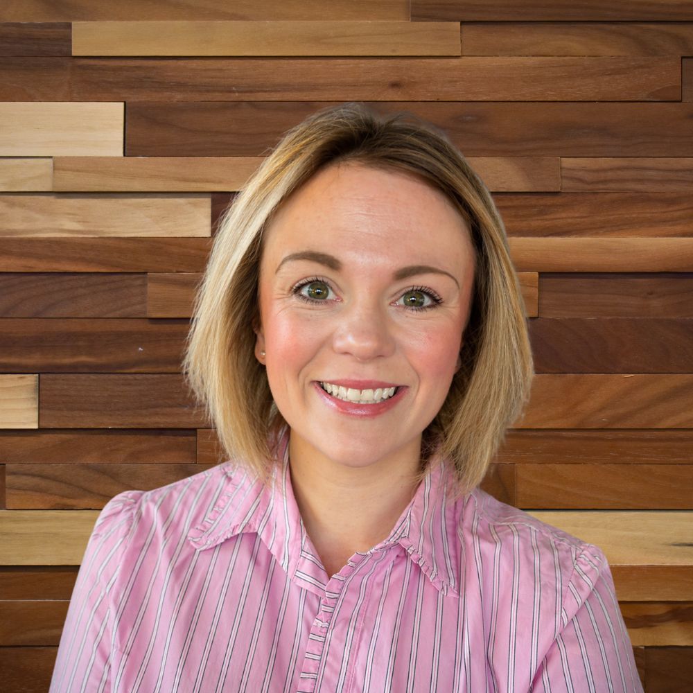 Smiling woman with short blonde hair wearing a pink striped shirt, standing in front of a wooden paneled wall.
